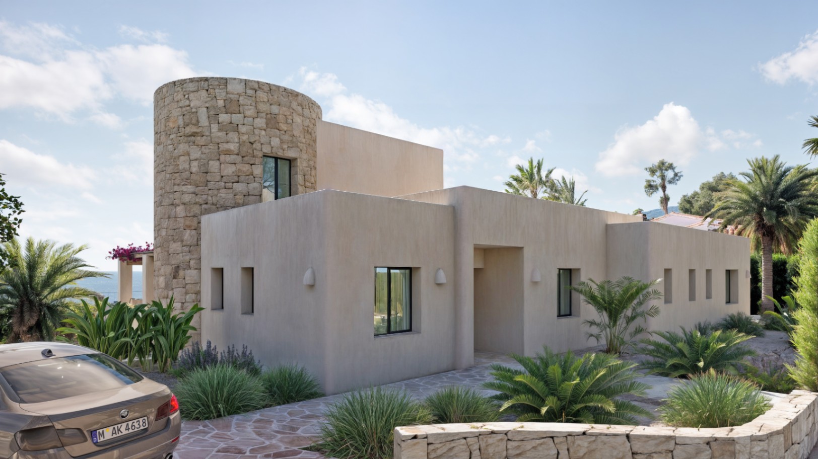 Fachada de estilo ibicenco con torre cilíndrica de piedra natural, carpintería de aluminio negro y jardín mediterráneo con pavimentación de piedra irregular.