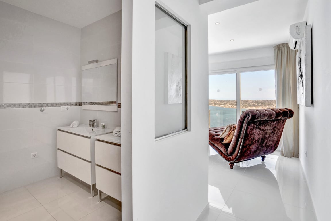 Baño en suite con lavabo doble y espejo con cenefa de mosaico, conectado visualmente a un dormitorio con chaise longue y vistas panorámicas al mar.