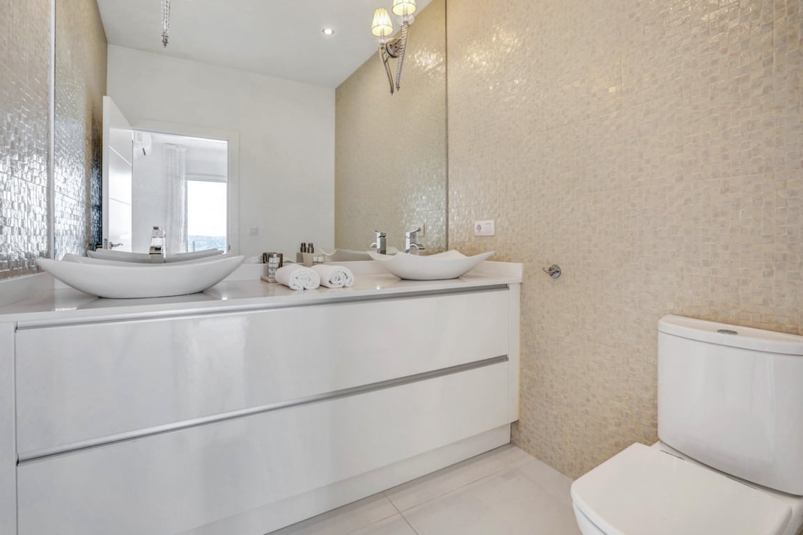Baño moderno con lavabo de doble seno sobre mueble blanco brillante, pared revestida en mosaico beige y gran espejo con apliques clásicos.