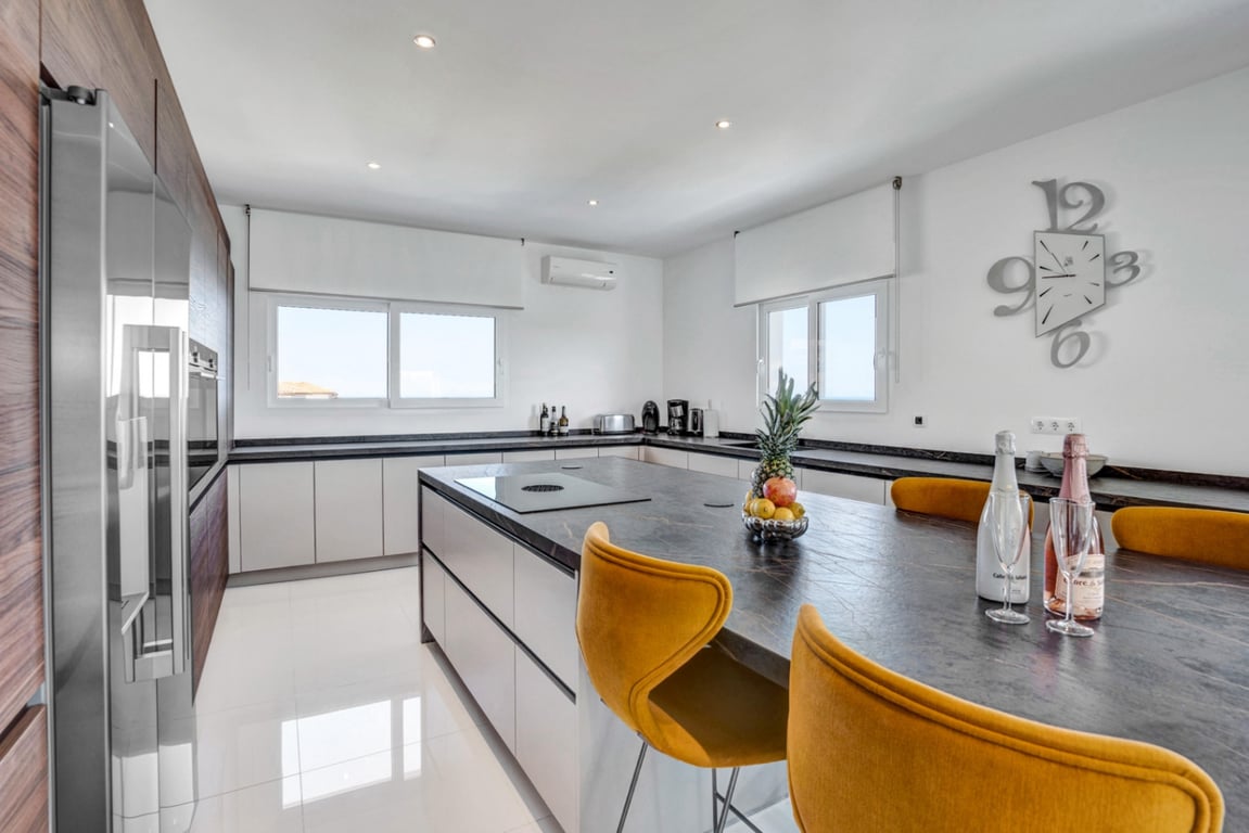 Cocina de diseño con isla central de piedra oscura, zona de desayuno con taburetes mostaza y armarios empotrados en madera y blanco sobre suelos brillantes.
