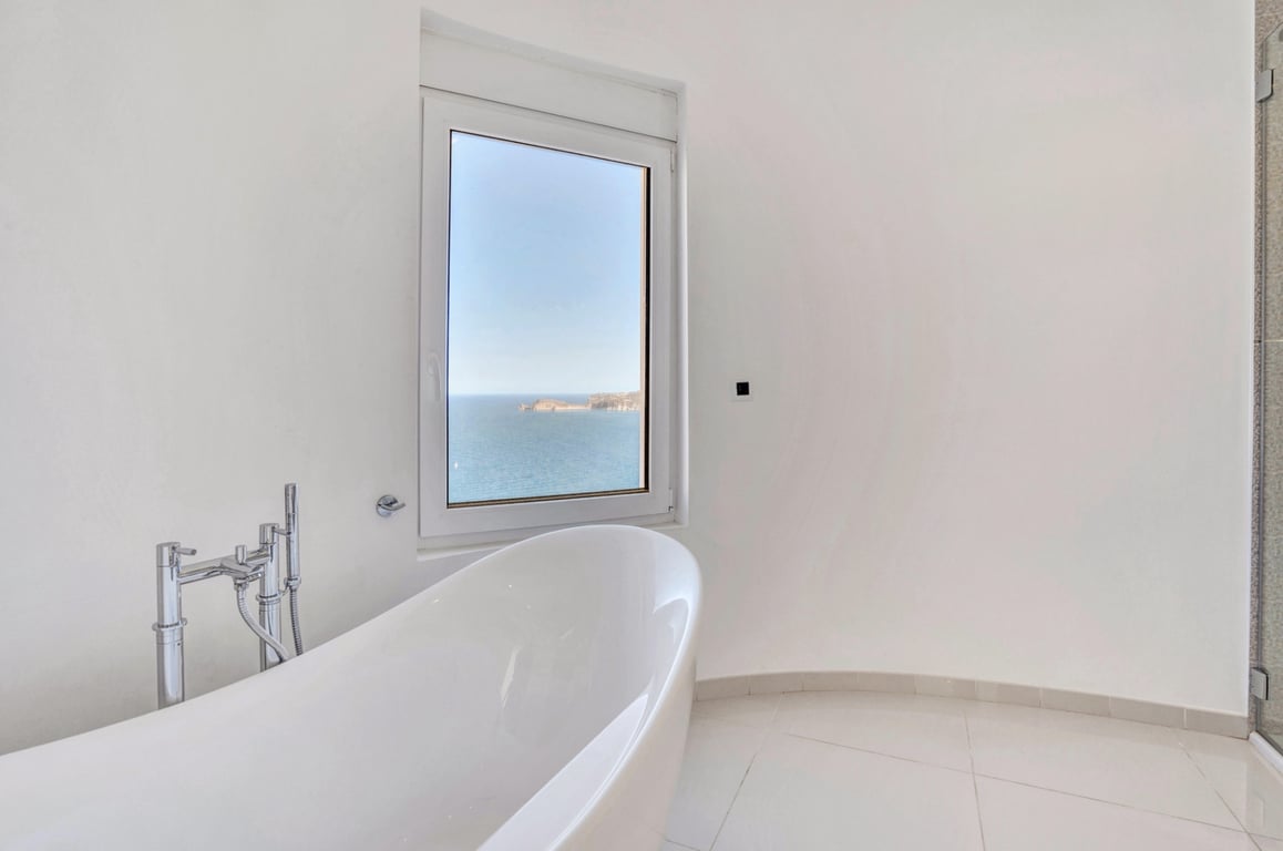 Cuarto de baño minimalista con bañera exenta blanca y grifería cromada, orientado hacia un ventanal con vistas panorámicas al mar Mediterráneo.