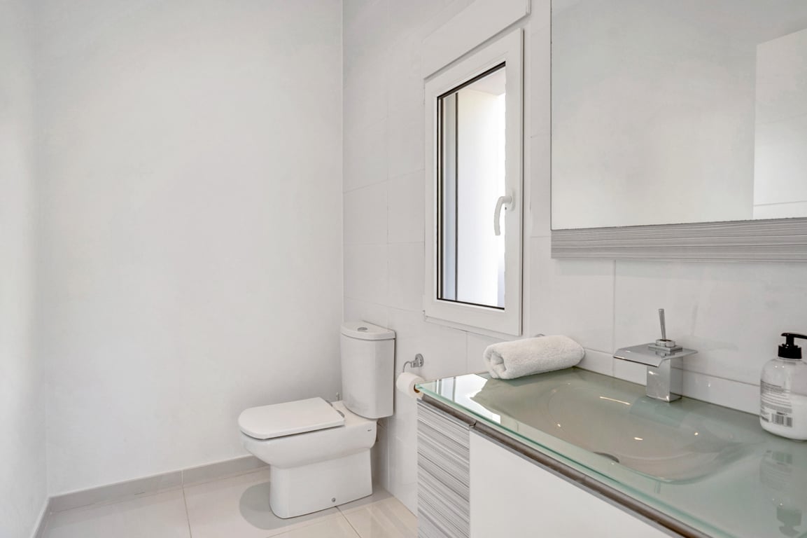 Cuarto de baño moderno con encimera de cristal translúcido, grifería cromada y paredes alicatadas en blanco con ventana oscilobatiente.
