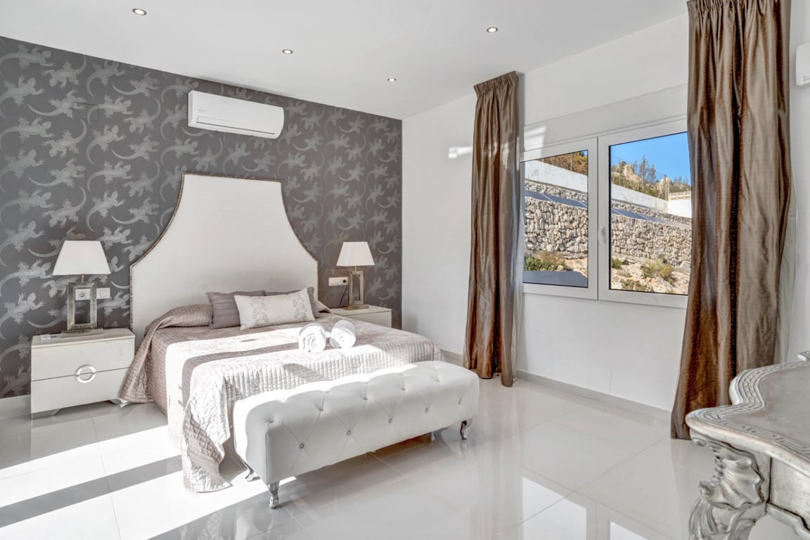 Dormitorio suite con pared de papel pintado gris, cama con cabecero blanco, aire acondicionado y ventanal con vistas al entorno rocoso de la parcela.
