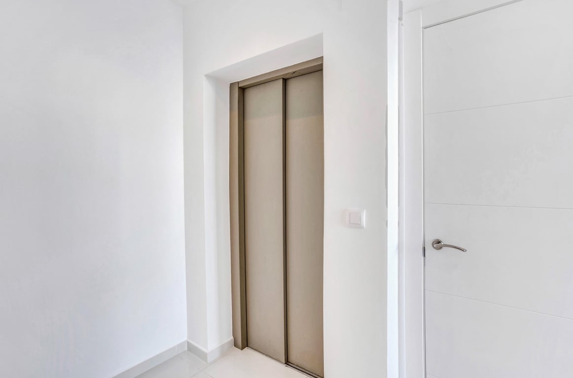 El ascensor privado con puertas metálicas en tono bronce se integra en un pasillo de diseño minimalista con paredes blancas y carpintería a juego.