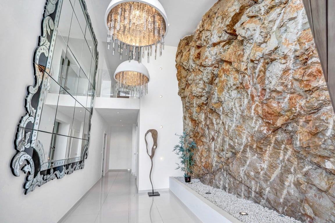 Pasillo de doble altura con una pared de roca natural integrada, lámparas de cristal de diseño y un gran espejo veneciano sobre suelos de gres pulido.