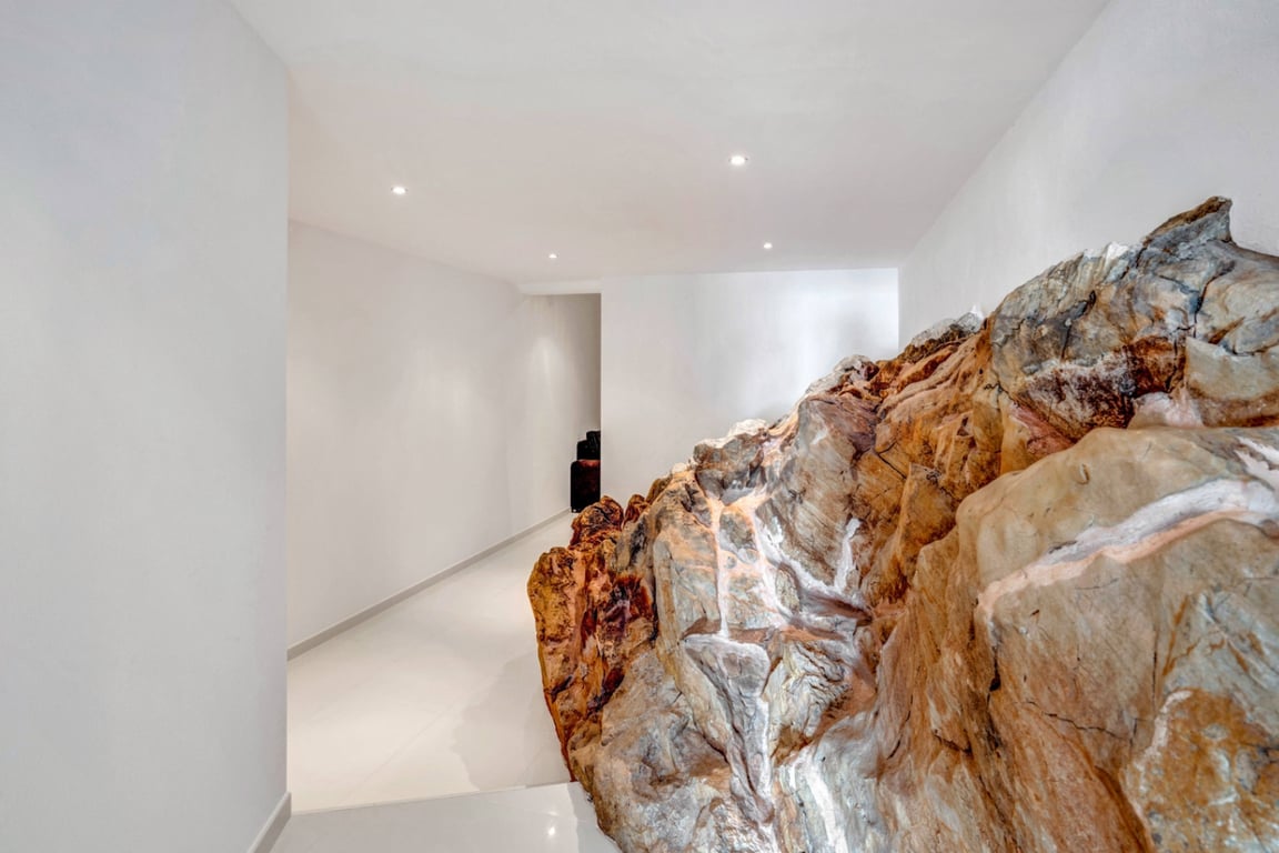 Pasillo interior con formación rocosa natural integrada en la arquitectura, paredes blancas minimalistas y luminarias empotradas en el techo.
