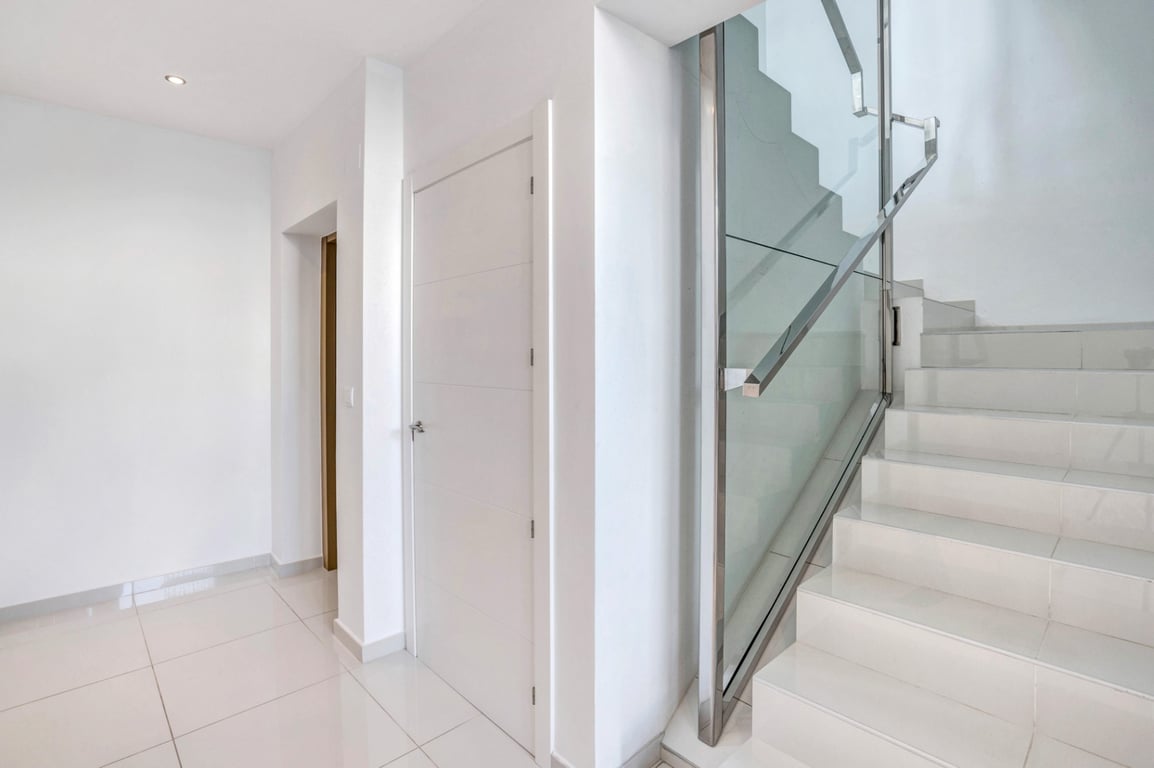 Pasillo interior con suelos cerámicos blancos y escalera de diseño moderno con barandilla de cristal y acero inoxidable.