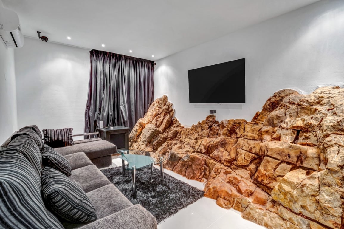 Sala de cine en casa con roca natural integrada en la pared, sofá gris, mesa de cristal y televisión de gran formato bajo iluminación empotrada.