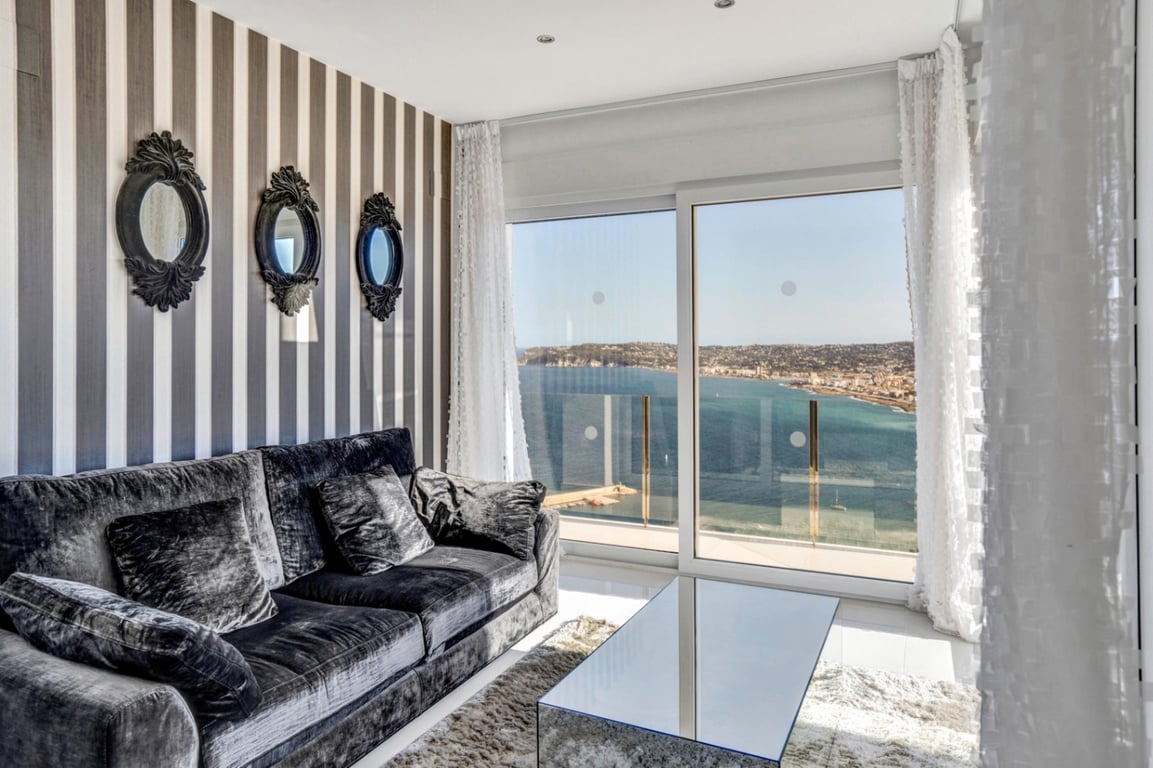 Sala de estar con sofá de terciopelo gris, papel tapiz de rayas y espejos ornamentados, ofreciendo vistas panorámicas al mar a través de grandes ventanales.