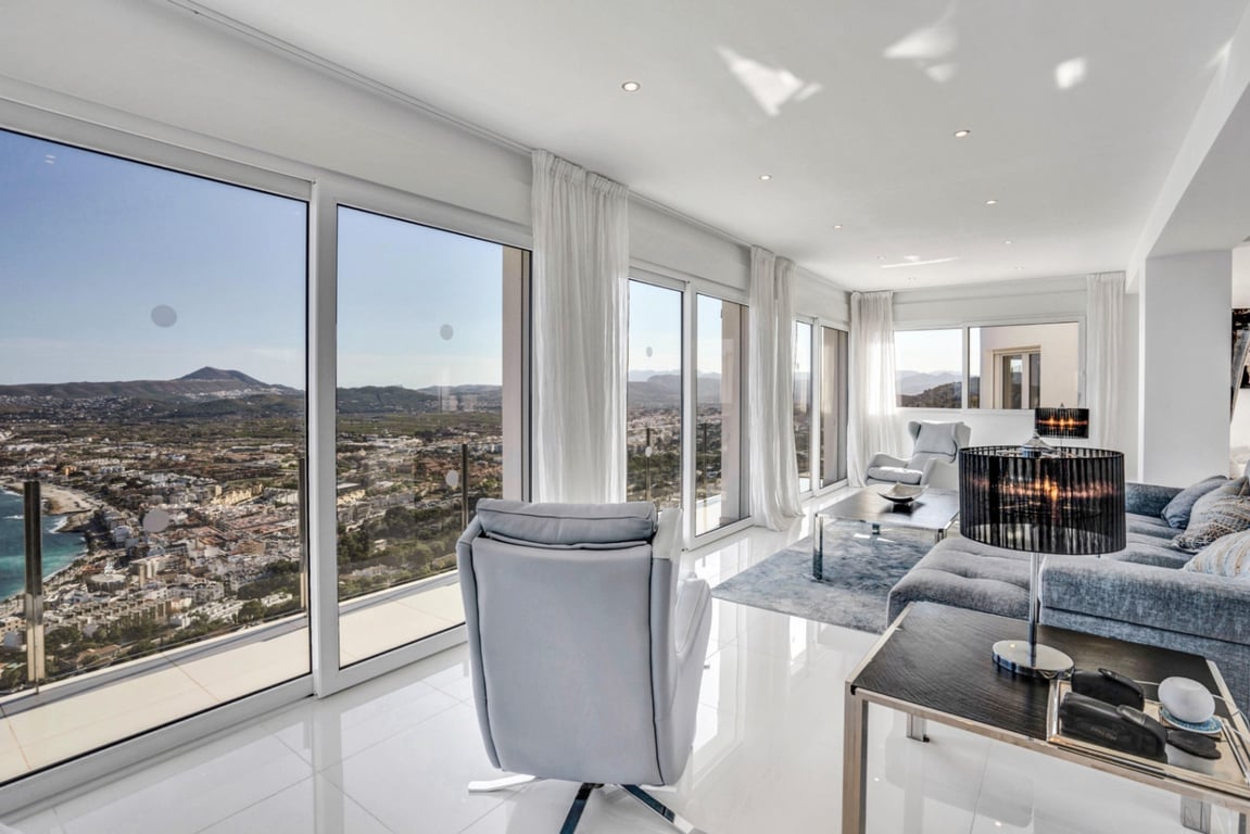 Salón luminoso con grandes ventanales de suelo a techo que ofrecen vistas panorámicas a la bahía de Jávea y las montañas, con suelos de gres blanco y diseño moderno.