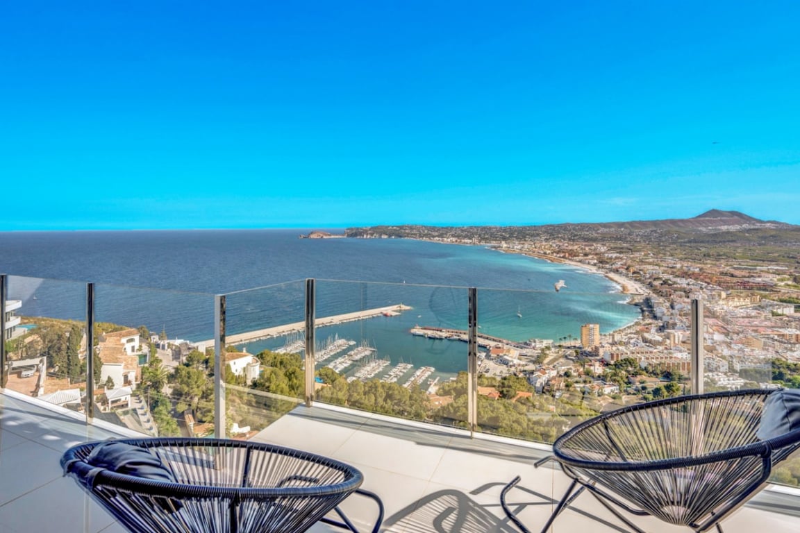 Terraza con barandilla de cristal y sillas Acapulco negras que ofrece una vista panorámica del puerto de Jávea y la bahía mediterránea.