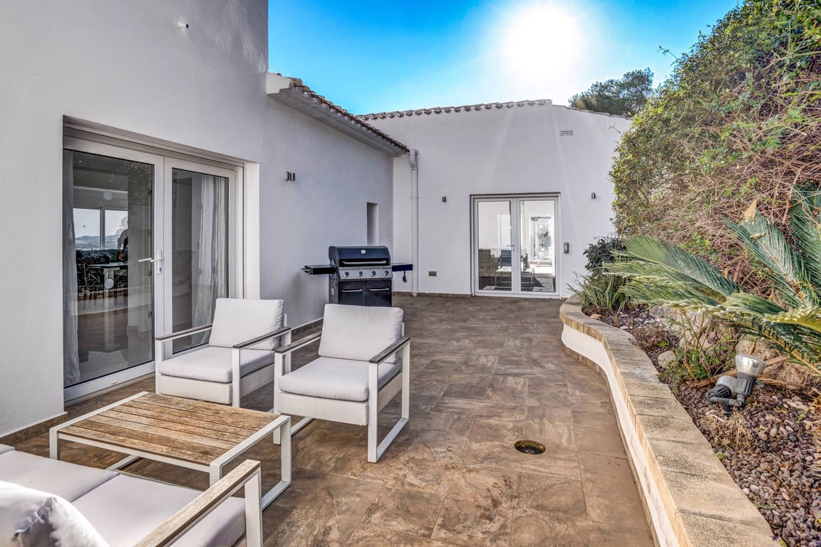 Terraza exterior con suelos de gres, zona de estar con sillones modernos, barbacoa y jardineras curvas con vegetación mediterránea.