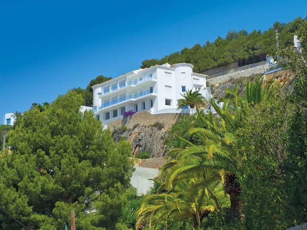 Vista exterior de la villa blanca integrada en la roca de La Corona, con múltiples niveles de terrazas, barandillas de cristal y rodeada de pinos y palmeras.
