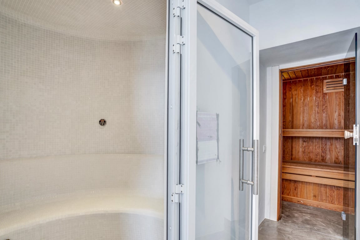 Zona de bienestar con baño turco revestido en mosaico blanco y sauna finlandesa de madera natural, integrados en el área de spa de la villa.