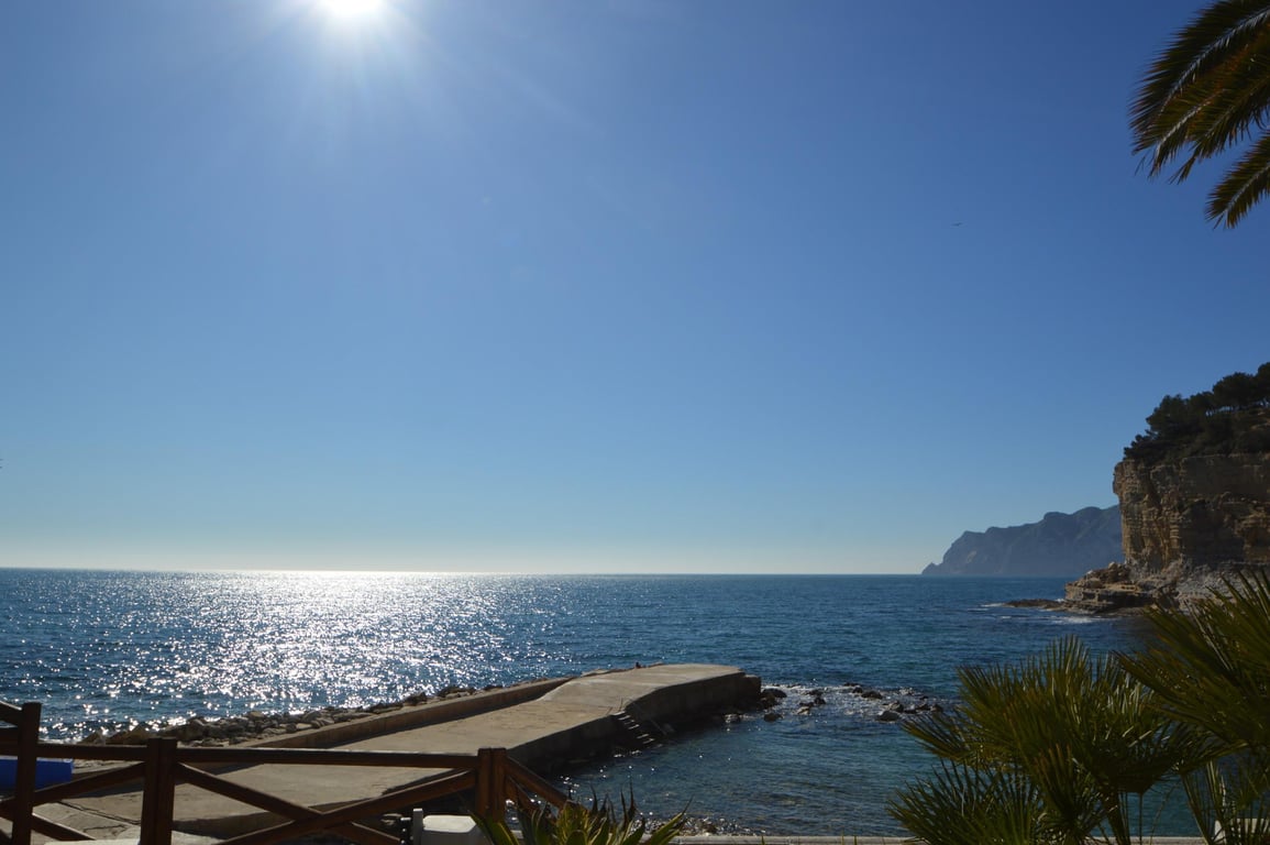 Vista de Cala Advocat con su espigón de hormigón y aguas cristalinas. El entorno destaca por sus acantilados rocosos y vegetación mediterránea bajo el sol.