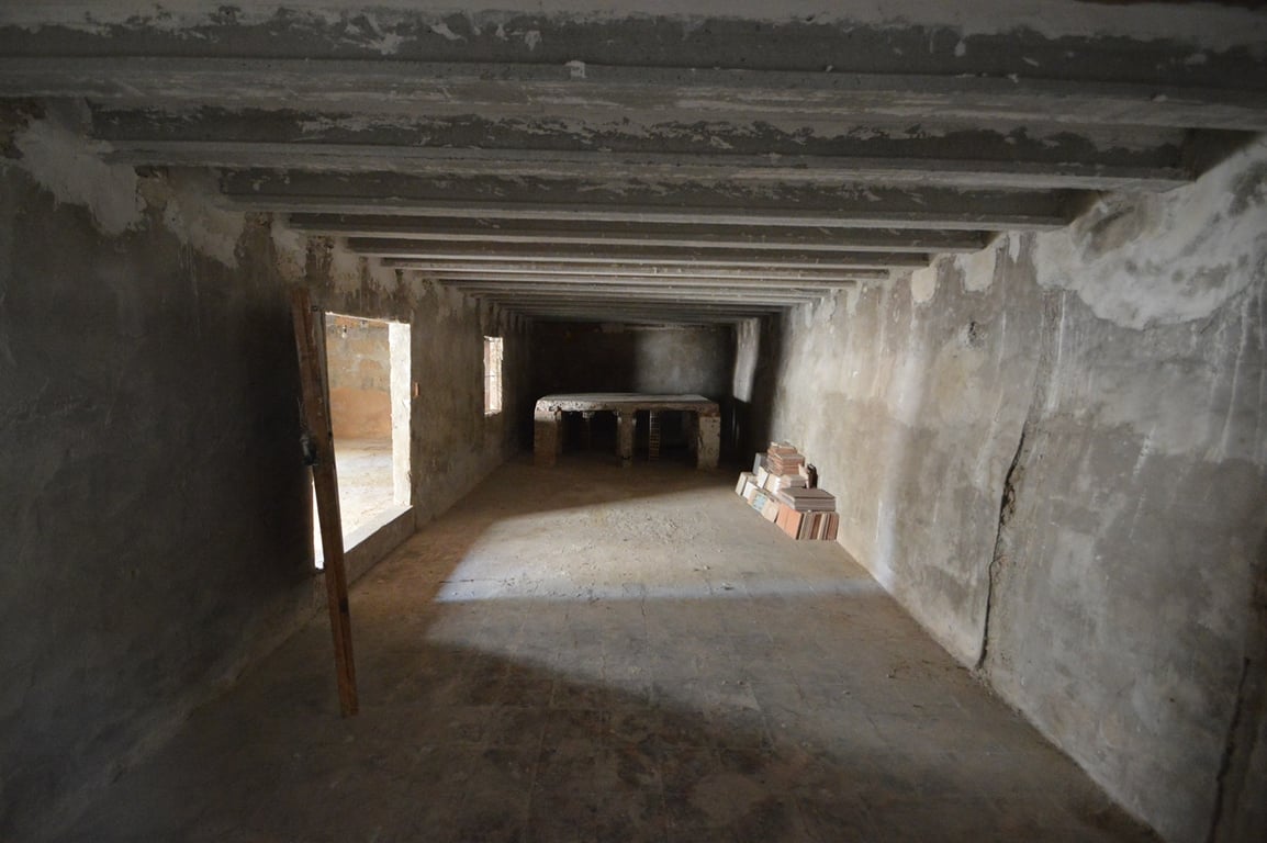 Amplio espacio diáfano en bruto con vigas de hormigón a la vista, paredes de piedra y una plataforma de obra al fondo, ideal para almacenamiento o taller.