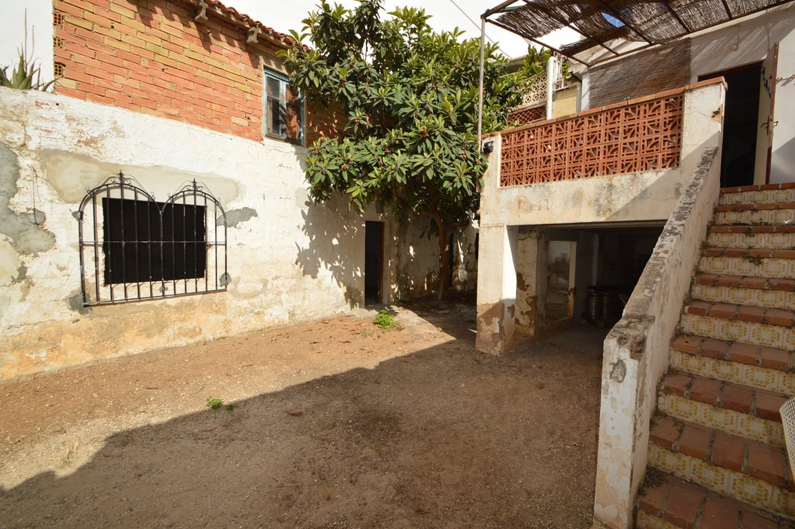 Patio interior rústico con suelo de tierra, gran níspero y escalera exterior de azulejos que sube a una terraza con pérgola de cañizo y barandilla cerámica.