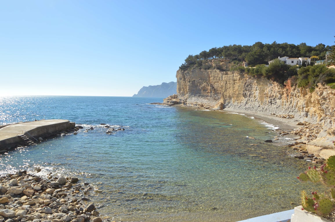 Vista de Cala Advocat con aguas cristalinas, acantilados de piedra caliza y un pequeño embarcadero de hormigón bajo el cielo despejado de la costa de Benissa.