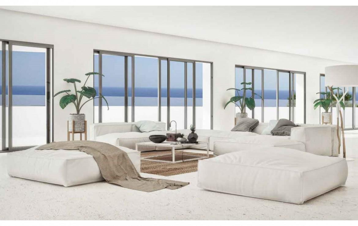 Salón minimalista con sofás modulares blancos, suelo de piedra clara y amplios ventanales con marcos oscuros que ofrecen vistas directas al mar Mediterráneo.