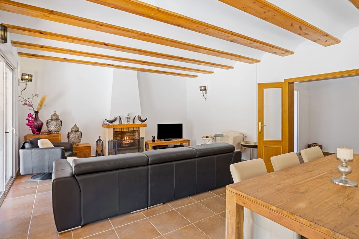 Amplio salón comedor con vigas de madera vista, suelos cerámicos y una chimenea central, conectado directamente con la terraza exterior.