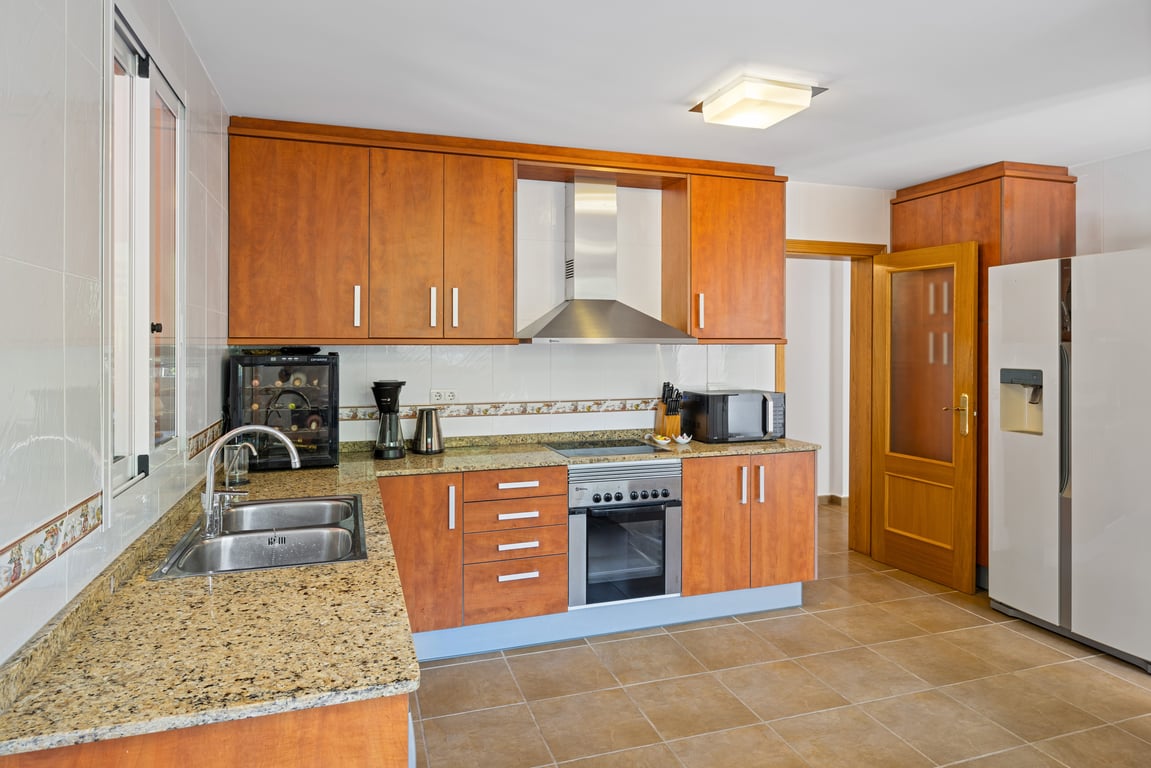 Cocina funcional en forma de L con armarios de madera, encimeras de granito, frigorífico americano y electrodomésticos integrados, incluyendo una vinoteca compacta.
