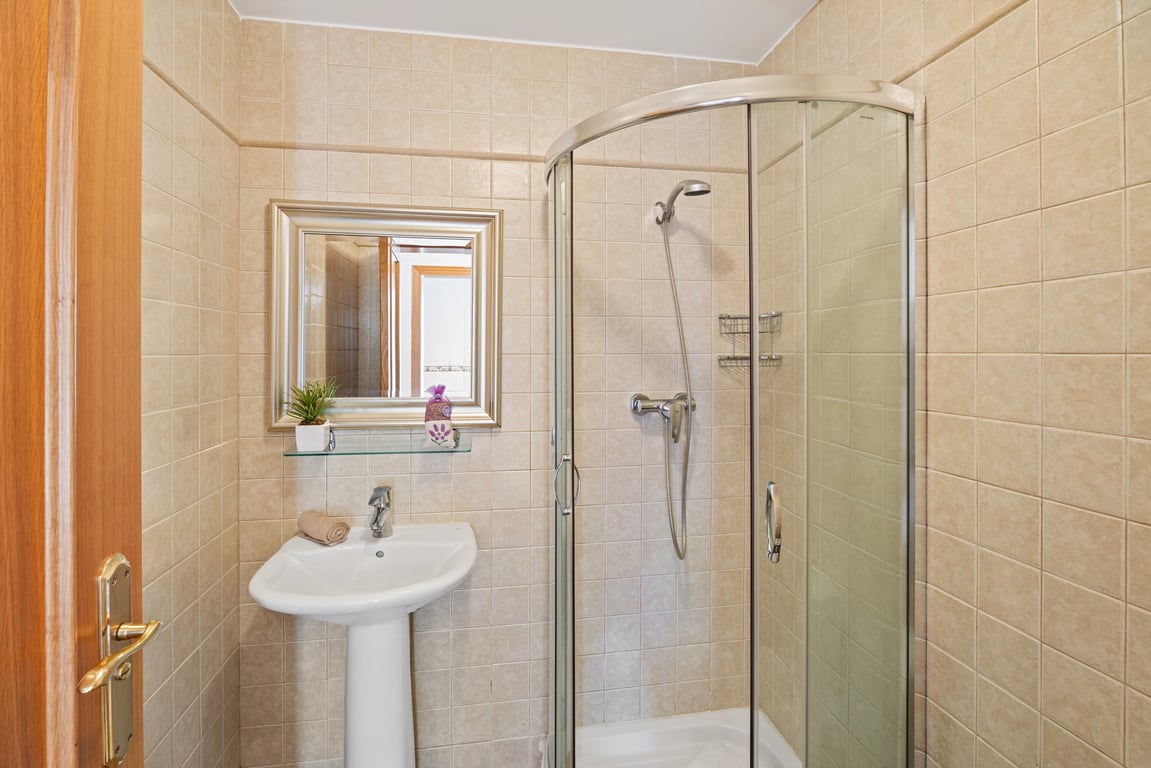 Cuarto de baño funcional con ducha de esquina de cristal curvo, lavabo de pedestal blanco y paredes revestidas con azulejos cerámicos en tonos beige.