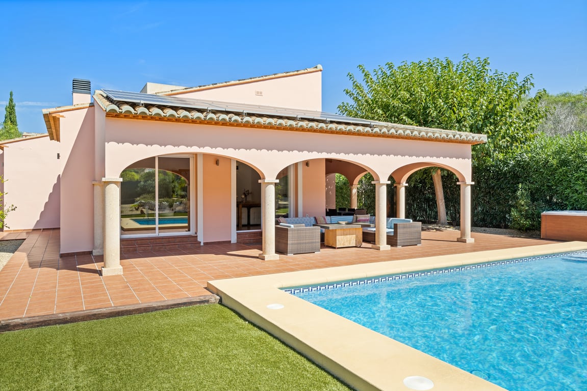 Exterior de la villa con piscina de mosaico, amplia naya arqueada con columnas de piedra y paneles solares integrados en el tejado, rodeada de césped artificial.