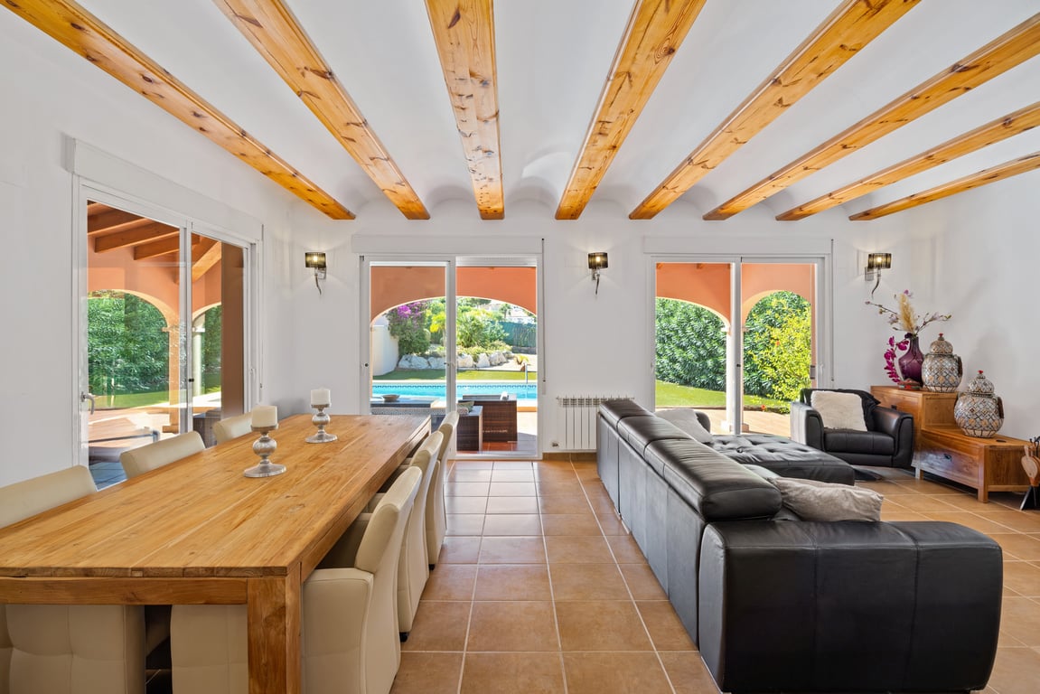 Salón-comedor luminoso con techos de vigas de madera, suelos de gres y amplios ventanales correderos que conectan directamente con la naya y la zona de la piscina.