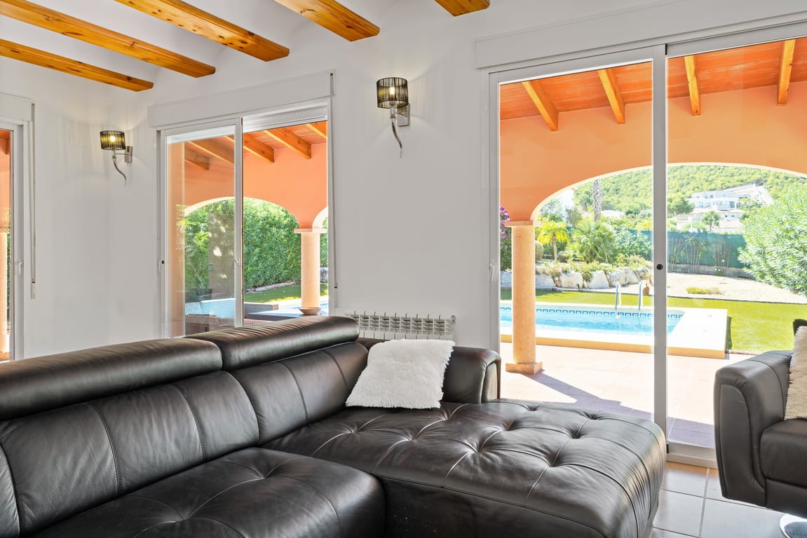 Salón luminoso con vigas de madera vistas, sofá de cuero negro y acceso directo a la naya y piscina a través de amplias puertas correderas de cristal.