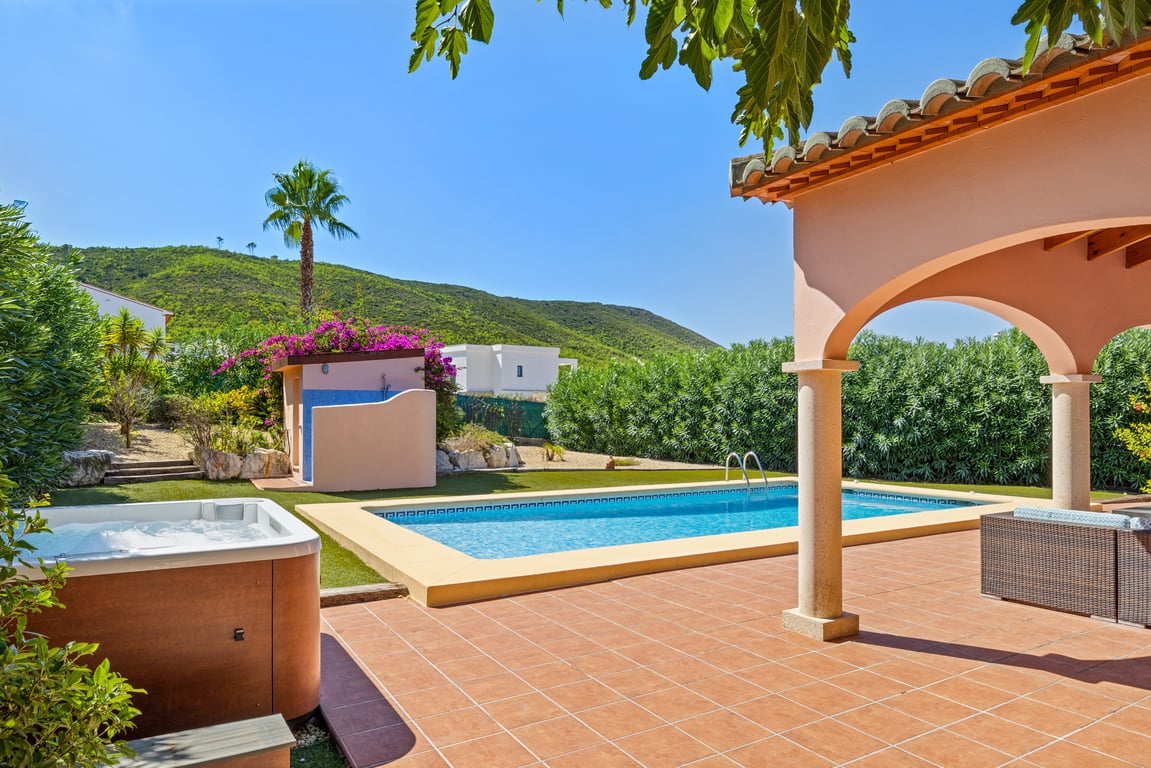 Terraza exterior con piscina de mosaico, jacuzzi privado y naya arqueada con columnas de piedra, todo con vistas despejadas a las montañas de Jávea.