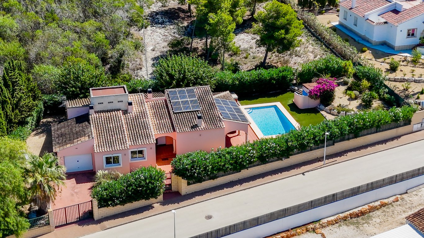 Vista aérea de la villa con tejado de teja, paneles solares y piscina privada rodeada de césped artificial y setos, ubicada en una parcela vallada junto a zona verde.