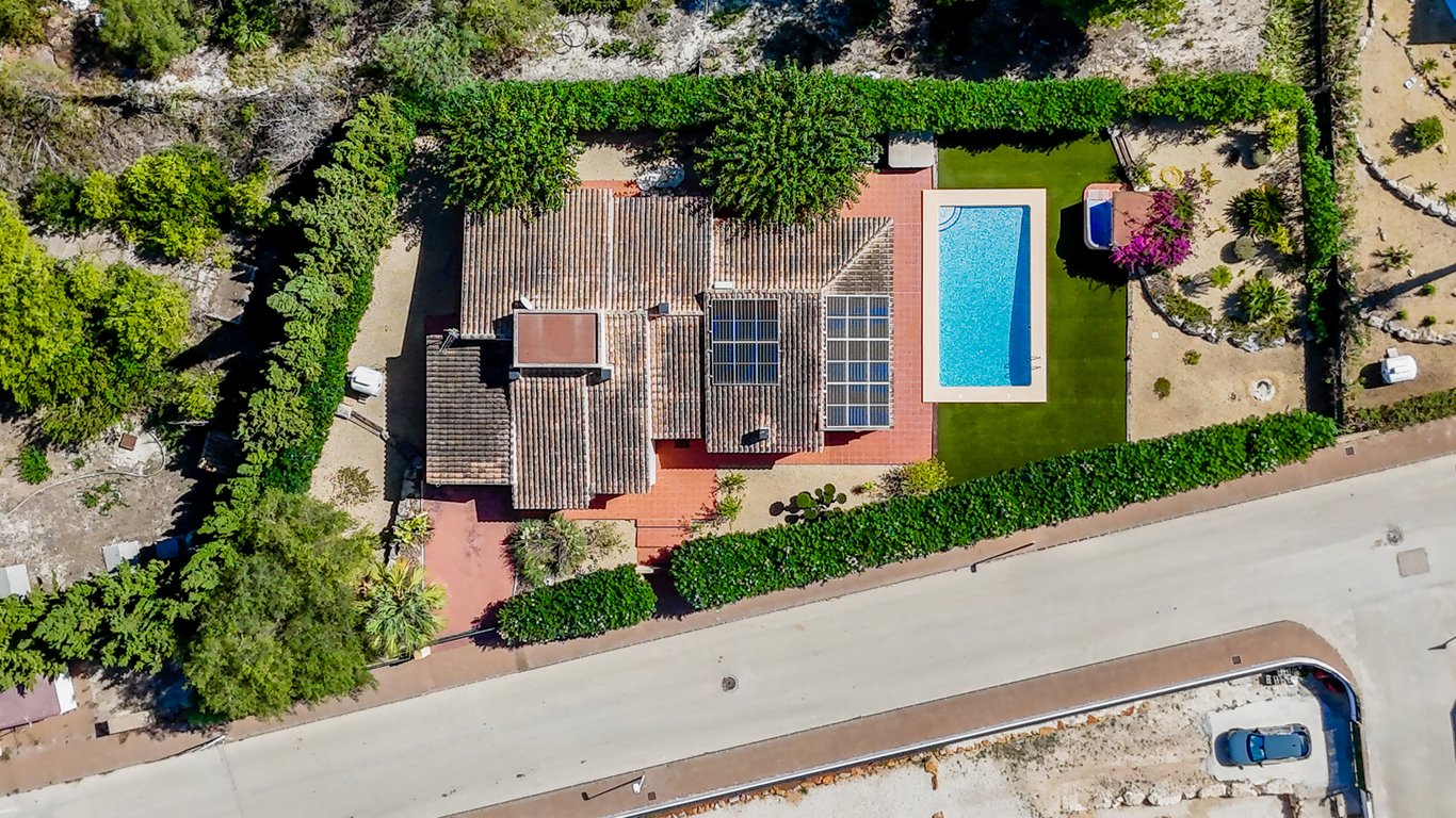 Vista aérea de la villa que muestra la distribución de la parcela, la piscina rectangular, los paneles solares en el tejado y el jardín perimetral con setos altos.