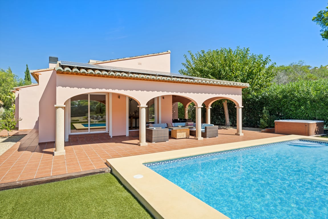 Vista exterior con piscina de mosaico, naya de arcos sostenida por columnas de piedra y paneles solares. La terraza de terracota incluye zona de estar y un jacuzzi lateral.