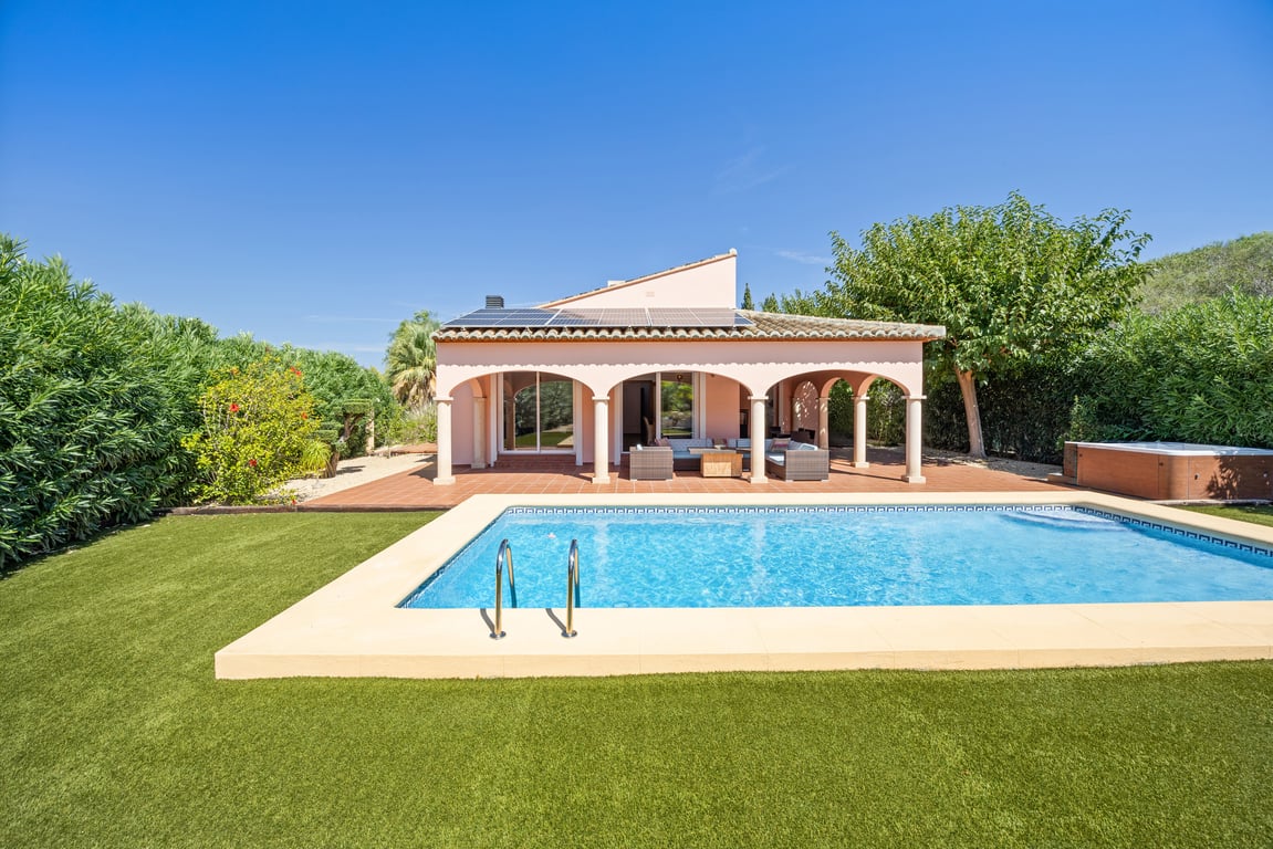 Vista exterior de la villa con piscina de mosaico, césped artificial y amplia naya arqueada. Destacan los paneles solares en el tejado y el jacuzzi lateral.