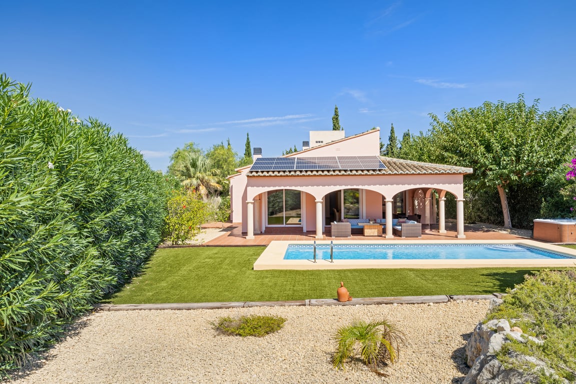 Vista exterior de la villa con piscina privada, naya arqueada y paneles solares integrados en el tejado, rodeada de un jardín de bajo mantenimiento con setos altos.