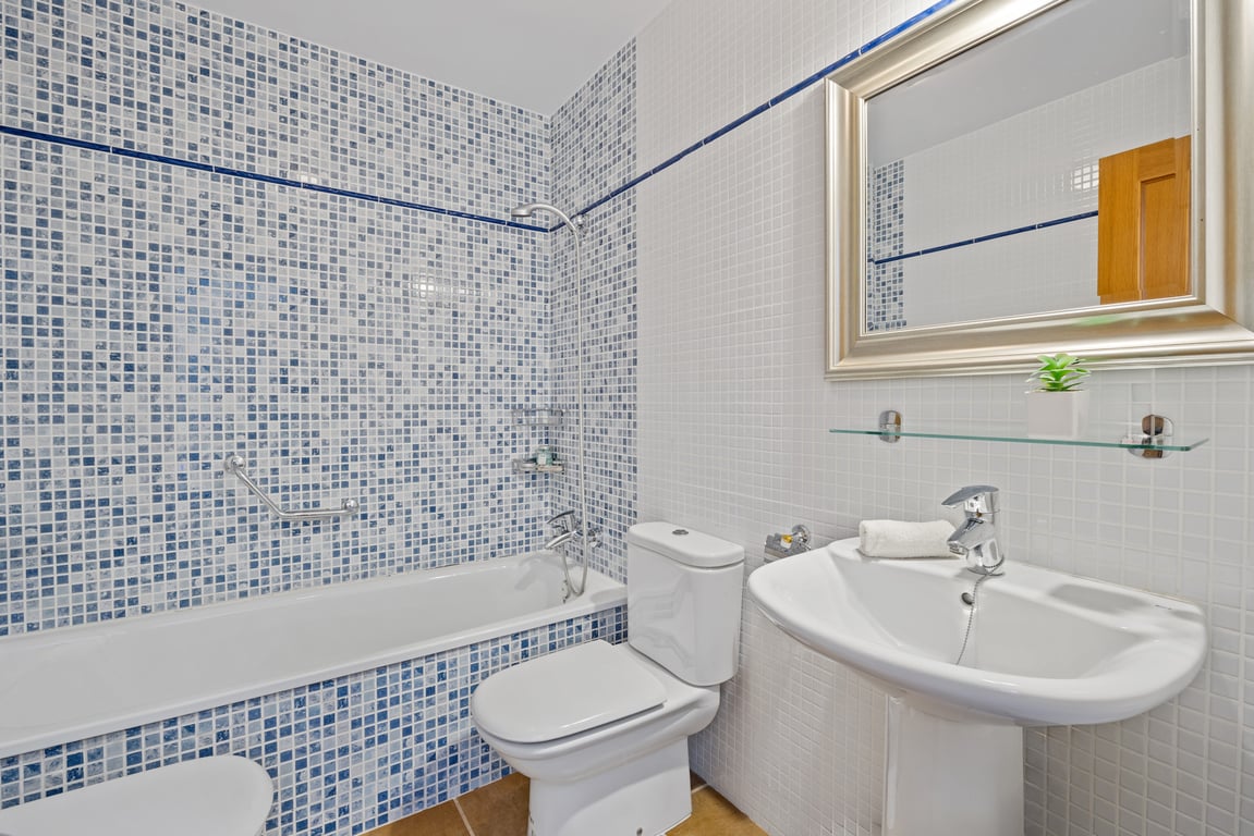 Cuarto de baño con bañera integrada, paredes revestidas de gresite azul y blanco, lavabo de pedestal blanco y espejo con marco plateado.