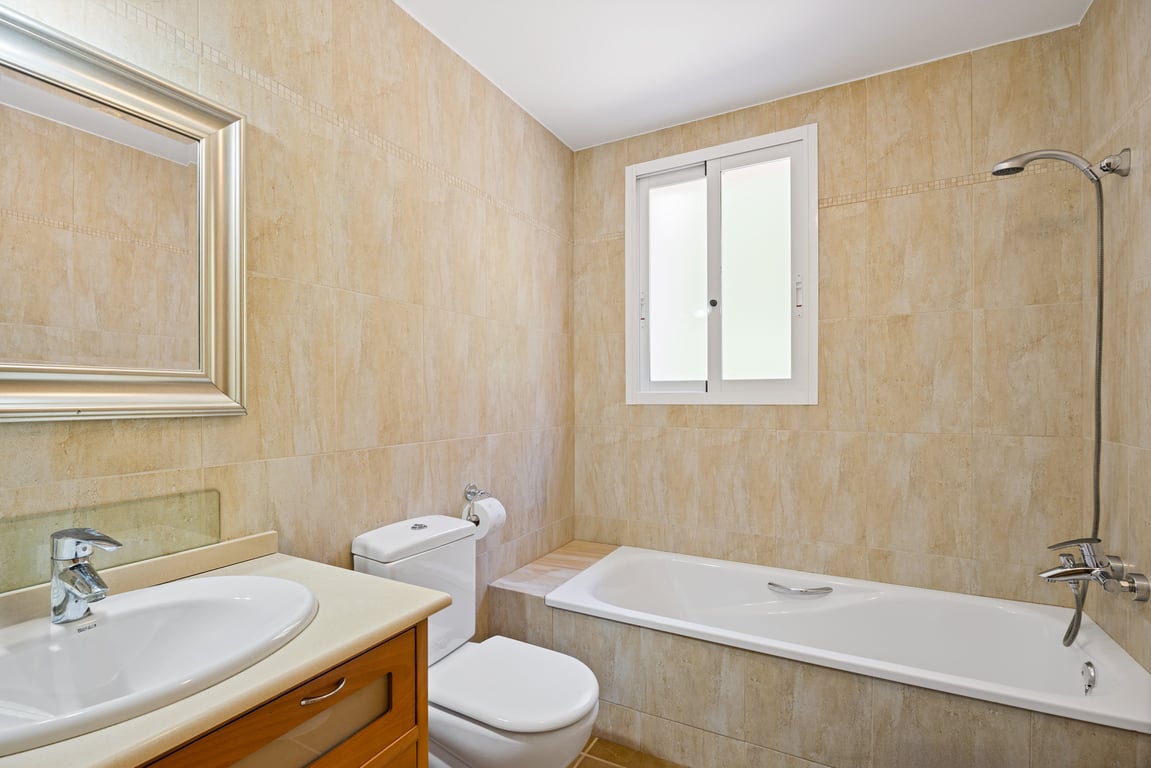 Cuarto de baño con paredes alicatadas en tonos beige, bañera encastrada con ducha, lavabo con mueble de madera y espejo de marco plateado.
