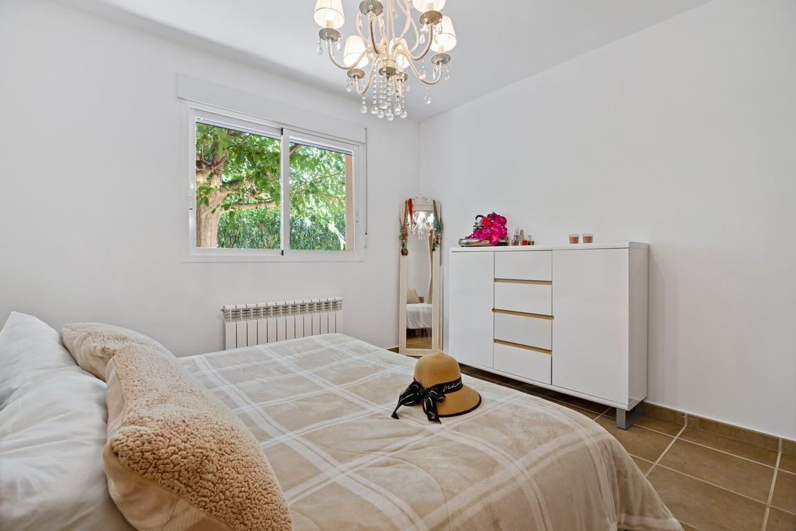 Dormitorio luminoso con paredes blancas, suelo cerámico, cómoda moderna, lámpara de araña clásica y ventana con vistas al jardín y radiador.