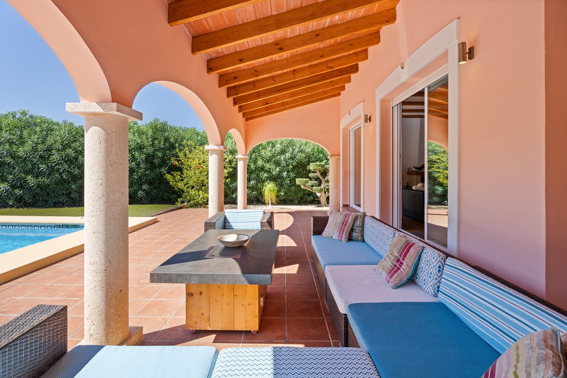Naya cubierta con vigas de madera vista, columnas de piedra tosca y zona de estar exterior con vistas directas a la piscina y al jardín.