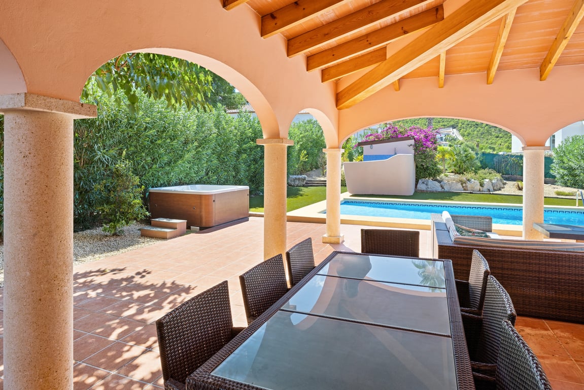 Naya cubierta con vigas de madera y arcos sobre columnas de piedra, con vistas a la piscina de mosaico, el jacuzzi exterior y el jardín mediterráneo.