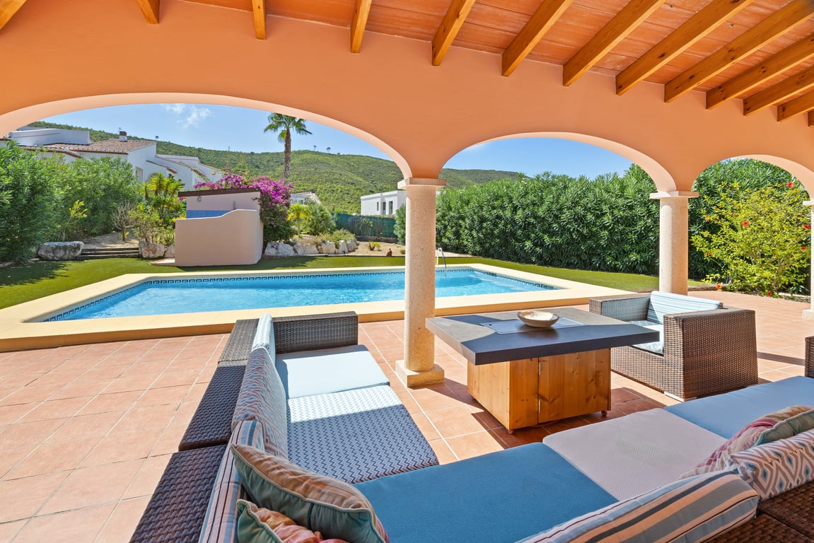 Naya cubierta con vigas de madera y arcos, zona de estar con sofás de mimbre frente a la piscina de mosaico y vistas a la montaña.
