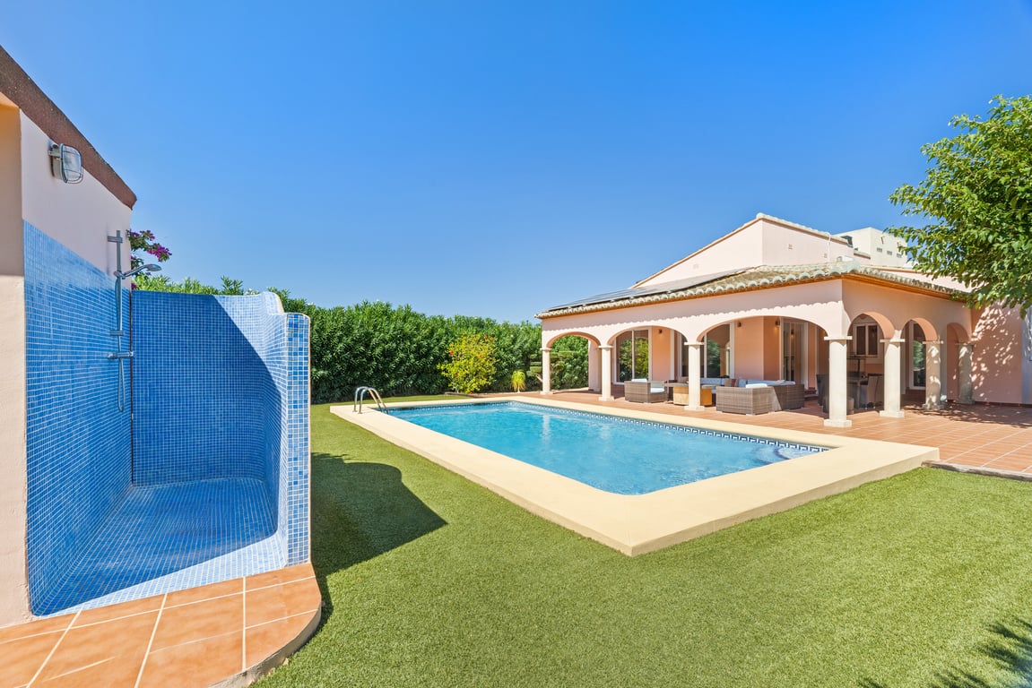 Piscina de mosaico azul rodeada de césped artificial, con ducha exterior revestida en gresite y una amplia naya arqueada con mobiliario de exterior.