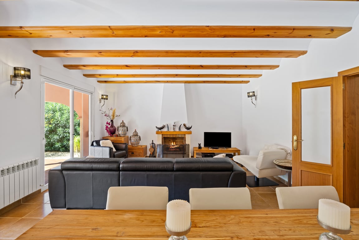 Salón luminoso con vigas de madera a la vista, chimenea central de obra y acceso directo a la naya exterior mediante puertas correderas.