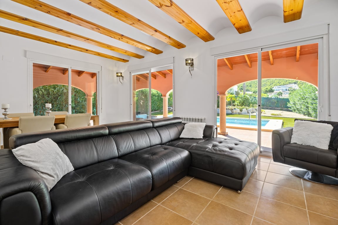 Salón luminoso con vigas de madera expuestas, sofá de cuero negro y grandes ventanales correderos con vistas directas a la piscina y la montaña.