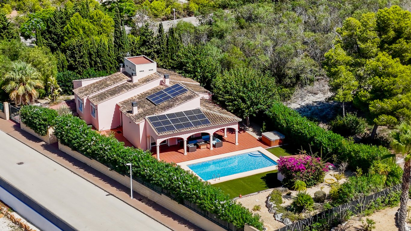 Vista aérea de la villa con paneles solares, naya arqueada frente a la piscina y jardín mediterráneo con jacuzzi y césped artificial.