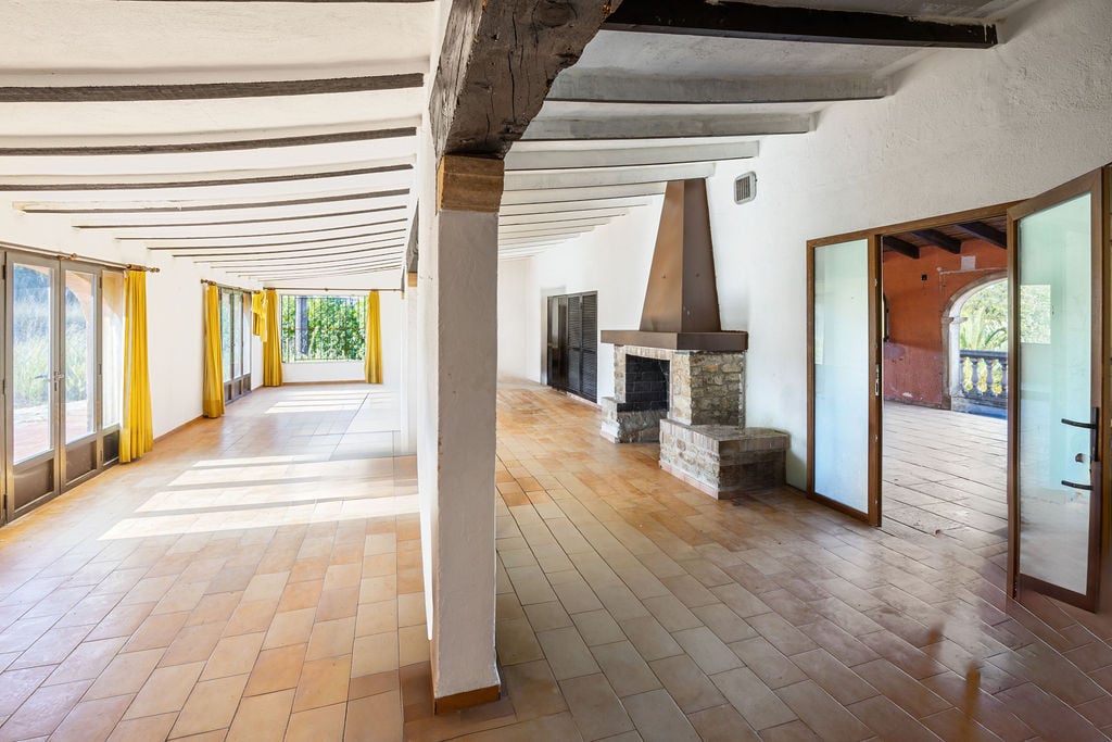 Amplio salón rústico con suelos de terracota, vigas de madera a la vista y una chimenea de piedra con campana metálica. La estancia destaca por su luz natural y techos altos.