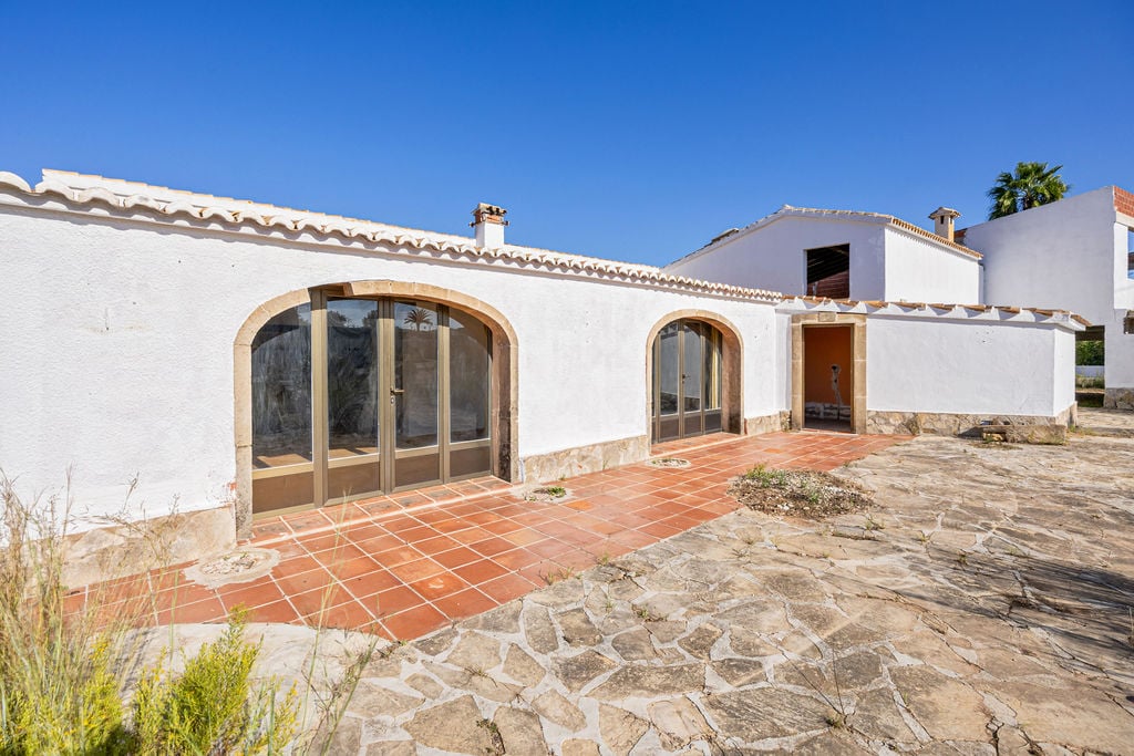 Fachada blanca con arcos de piedra y amplios ventanales. El exterior combina suelos de terracota con pavimentos de piedra natural en un entorno mediterráneo despejado.