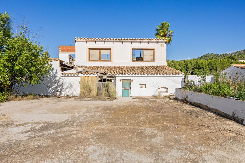 Fachada de construcción tradicional con muros blancos, carpintería de madera y techos de teja árabe sobre un amplio terreno nivelado rodeado de vegetación mediterránea.