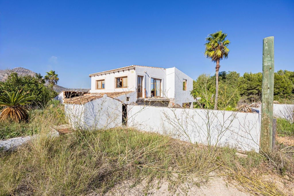Fachada de estilo rústico mediterráneo con muros blancos y cubiertas de teja árabe, rodeada de vegetación natural y palmeras bajo un cielo despejado.