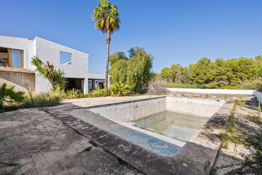 Piscina exterior con detalles de mosaico y amplia zona de terraza, rodeada de vegetación mediterránea y una edificación blanca de arquitectura contemporánea.